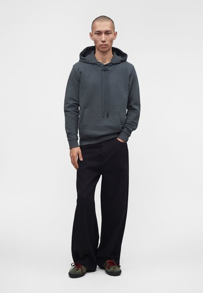Homme debout avec une main dans la poche, portant un sweat à capuche gris foncé, un pantalon large noir et des baskets vertes avec des lacets rouges, sur un fond uni.