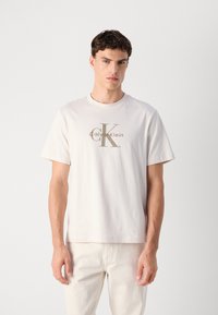 Lys beige bomulds t-shirt med rund halsudskæring, korte ærmer og et centralt logo-design i en mørkere beige nuance.