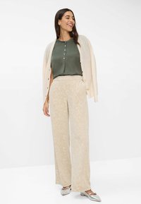 Crème cardigan, groene blouse met knopen, beige met patronen gevlekte broek en zilveren ballerina's. Losse pasvorm, zachte texturen en subtiele details op de broek.