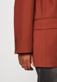 Gros plan sur la main d'une personne à côté de la manche d'un blazer couleur rouille avec quatre boutons, porté avec un pantalon gris foncé.