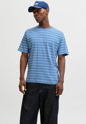 Man die een blauw-wit gestreept T-shirt draagt met tekst op de borst, donkere broek met wijde pijpen en een blauwe baseballpet, kijkt opzij.