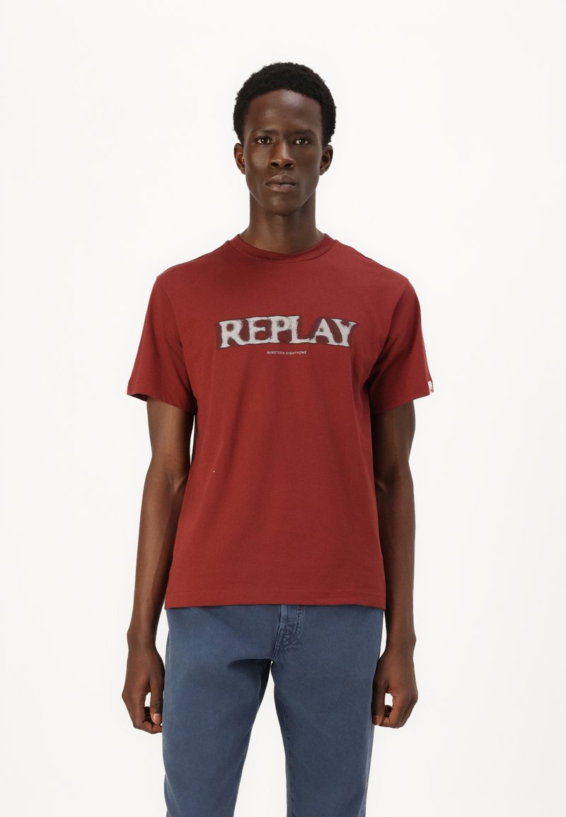 Camiseta de algodón roja con cuello redondo, que presenta un prominente logo gris "REPLAY" en el pecho. Manga corta y corte regular.