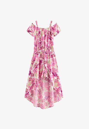 Ung kvinde iført en pink og beige floral off-shoulder kjole med høj-lav kant, stående barfodet mod en ensfarvet hvid baggrund.