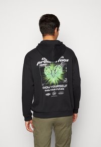 Sudadera negra con estampado gráfico en la parte posterior que presenta formas verdes y afiladas junto con texto. Fabricada en tejido suave, cuenta con un bolsillo tipo canguro y capucha con cordón.