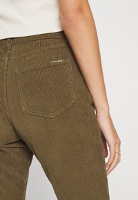 Gros plan sur une personne portant des pantalons en velours côtelé vert olive et un haut à côtes blanc, montrant la poche arrière et une partie du bras droit avec une bague.