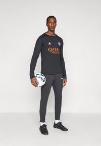 Μαύρη μπλούζα με μακριά μανίκια και πορτοκαλί κείμενο "QATAR AIRWAYS", λογότυπο στο στήθος; σκούρο γκρι αθλητικό παντελόνι; κρατάει μια λευκή και μπλε μπάλα ποδοσφαίρου.