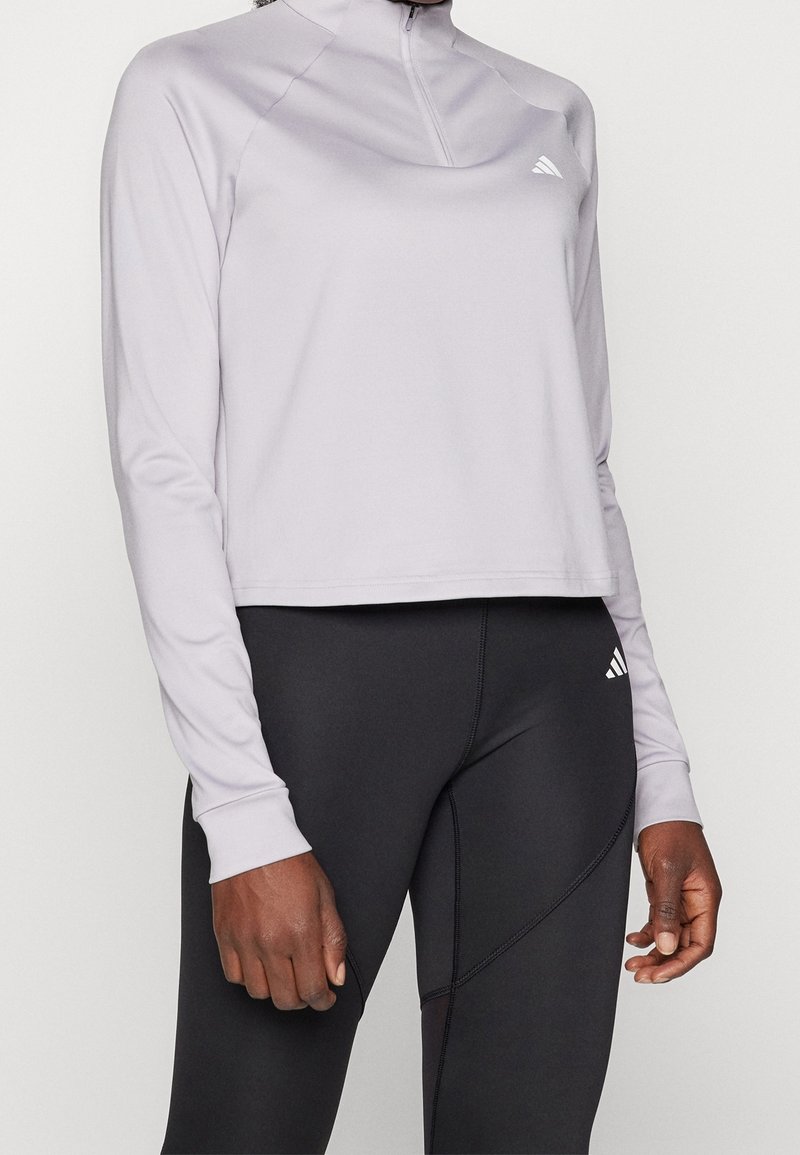 Vaaleanharmaa urheilupaita puolivetoketjulla ja pitkillä hihoilla, yhdistettynä istuviin mustiin leggingsseihin, joissa on ompelukoristeita ja yksityiskohtia.