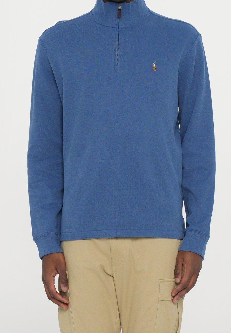 Homme portant un pull bleu à manches longues avec une fermeture éclair partielle et un pantalon cargo beige sur un fond uni.