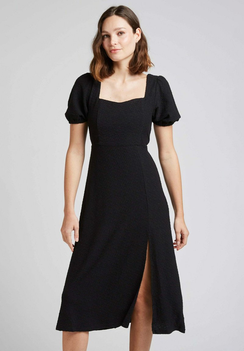 Robe noir midi avec décolleté carré, manches bouffantes, tissu texturé et une fente à l'avant, créant une silhouette ajustée.