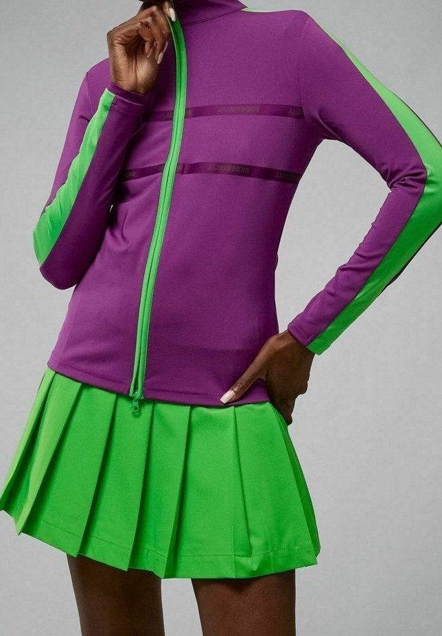 Personne portant une veste zippée violette et verte avec des bandes vertes sur les manches, associée à une jupe plissée vert vif, posant avec la main sur la hanche.