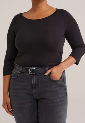 Vrouw die een zwart top met driekwart mouwen draagt, ingestopt in hoogwater vervaagde zwarte jeans met een zwarte riem, haar rechterhand in de zak.