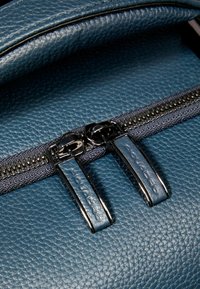 Mochila de cuero color azul verdoso con un acabado texturizado, que cuenta con una doble cremallera con tiradores metálicos y un detalle de cremallera de tela en contraste.