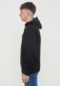 Svart hettegenser med snor, frontlomme og ribbede mansjetter. Båret med blå jeans, vist fra siden mot en ensfarget bakgrunn.