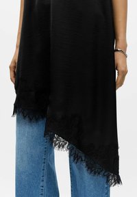 Sort asymmetrisk topp med teksturert finish, med intrikat svart blonderkant nederst, kombinert med lyseblå jeans.