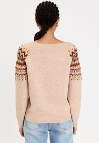 Woman with braided hair wearing a beige sweater with multicolored patterned sleeves and light blue denim jeans, viewed from behind.