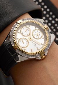 Montre en or et argent avec un cadran rond, trois sous-cadrans, des accents en cristal et un bracelet en caoutchouc noir. La marque "GUESS" est visible sur le cadran.