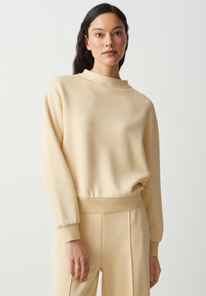 Frau mit langen dunklen Haaren, die einen beigen Pullover mit hohem Kragen und passende hochtaillierte Hosen trägt, steht vor einem schlichten Hintergrund.