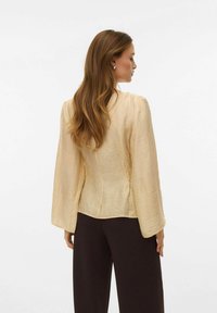 Blusa beige chiaro testurizzata con maniche lunghe svasate, caratterizzata da un corpetto aderente. Indossata con pantaloni marrone scuro.