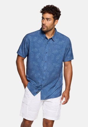 Hombre con camisa azul de manga corta con botones y estampado de hojas, y pantalones cortos cargo blancos, de pie con una mano en el bolsillo, mirando hacia la izquierda.