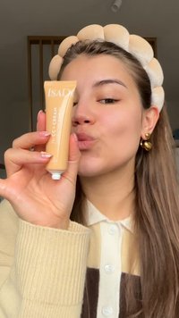 Tube beige de crème CC ISADORE tenue à la main, avec un texte blanc. La personne porte un pull côtelé beige et marron avec un col.