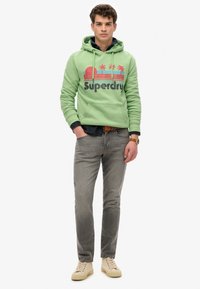 Sudadera verde con gráfico de palmera y atardecer, combinada con jeans grises y zapatillas beige. Cuenta con un bolsillo tipo canguro y capucha con cordón.