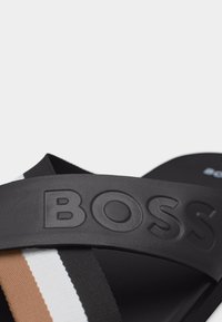 Μαύρη σαγιονάρα slide με ανασηκωμένο λογότυπο "BOSS" στο λουράκι και επικαλυπτόμενες ταινίες υφάσματος σε καφέ, λευκό και μαύρο από κάτω.