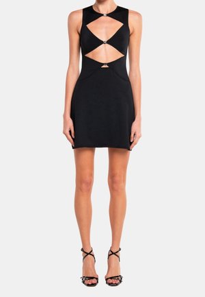 Robe noire sans manches avec détails découpés et accents argentés, silhouette ajustée, tissu lisse. Associée à des sandales à talons noires à brides.