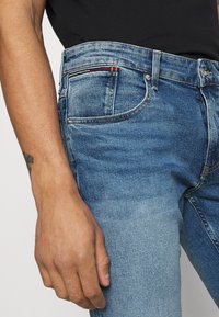 Siniset farkut, joissa on lievä kulutus, viisi taskua, kuparin värinen ompelu ja erottuva punainen ja keltainen yksityiskohta vyötäröllä.