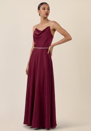 Frau trägt ein ärmelloses, bodenlanges Satinballkleid in Burgunderrot mit dünnen silbernen Trägern und einem silbernen Taillenband, steht mit einer Hand in der Hüfte.