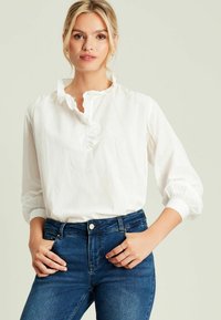 Weiße Bluse mit langen Ärmeln und rüschiertem Ausschnitt, mit elastischen Manschetten, kombiniert mit dunkelblauen hochtaillierten Jeans. Einfaches, sauberes Design.