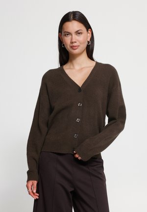 Femme aux longs cheveux foncés portant des boucles d'oreilles argentées en forme de créoles, un cardigan marron boutonné et un pantalon sombre sur un fond uni.
