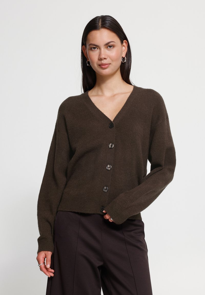 Femme aux longs cheveux foncés portant des boucles d'oreilles argentées en forme de créoles, un cardigan marron boutonné et un pantalon sombre sur un fond uni.
