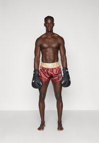 Pantalones de boxeo en satén color burdeos con una cinturilla elástica en dorado, que presenta un logo. Guantes de boxeo negros con detalles en blanco, pies descalzos.