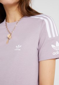 Ljuslila kortärmad skjorta med tre vita ränder på axlarna och vit Adidas-logga på bröstet och ärmen. Slät, mjuk tyg.