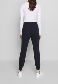 Vrouw die met haar rug naar de camera staat, gekleed in een witte langemouwen top, zwarte joggers met elastische tailleband en witte sneakers, tegen een effen achtergrond.