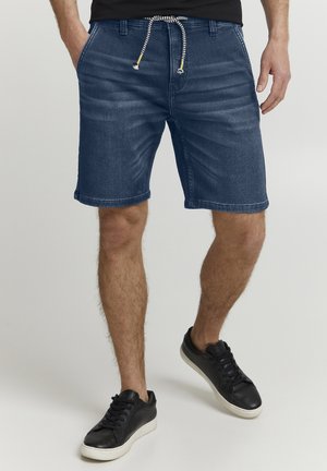 Shorts en denim bleu foncé avec une coupe décontractée, présentant une taille élastiquée dotée d'un motif noir et blanc, et des ourlets repliés. Baskets noires.