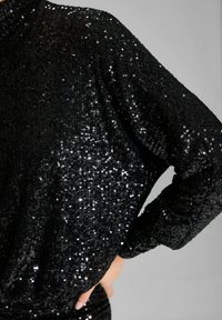 Top negro con lentejuelas, de cuello alto, mangas largas y un brillo texturizado creado por el solapamiento de lentejuelas para un efecto reflectante.