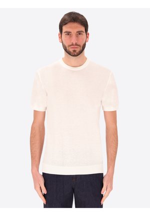 Uomo con capelli corti scuri e barba, che indossa una maglietta a maniche corte bianca semplice e pantaloni scuri, in piedi di fronte su sfondo bianco.