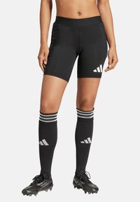 Schwarze Sportshorts aus glattem Stoff, mit drei weißen diagonalen Streifen an der Seite. Kombiniert mit passenden knielangen Socken und Stollenschuhen.