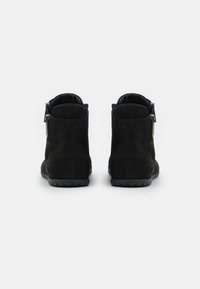 Pár černých semišových kotníkových bot s bočními zipy, viděný ze zadní strany na čistém bílém pozadí.