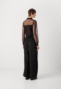 Blouse noire transparente avec un motif à pois, manches longues et un dos en dentelle texturée. Associée à un pantalon noir à jambes larges et bien ajusté.