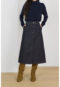 Falda A-line de mezclilla en azul oscuro con bolsillos frontales, cierre de cremallera y costuras naranjas, combinada con botas de ante marrón hasta la rodilla.