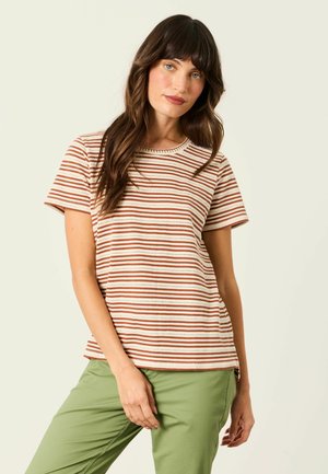 Junge Frau mit langen braunen Haaren, die ein cremefarbenes und rostfarben gestreiftes kurzärmeliges Shirt und hellgrüne Hose trägt, steht vor einem schlichten Hintergrund.