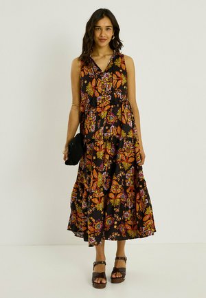 Femme portant une robe noire fleurie sans manches, tenant une pochette noire et portant des sandales marron à plateforme, debout devant un fond uni.