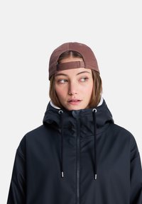 Μαύρο αδιάβροχο μπουφάν με ψηλό γιακά και ρυθμιζόμενα κορδόνια, σε συνδυασμό με καφέ υφασμάτινο καπέλο με λουράκι πίσω.