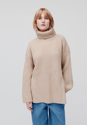 Beżowy sweter dziany z wysokim kołnierzem, z prążkowanym wzorem i opadającymi ramionami. W połączeniu z luźnymi niebieskimi dżinsami.