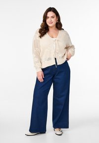 Beige öppenstickad cardigan med knytframsdetalj, ihop med vida mörkblå jeans. Enkel design, inga märkbara mönster eller accenter.