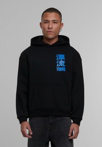 Sudadera negra con un bolsillo en el frente, con texto japonés en azul en el lado izquierdo. El material parece suave; la capucha y los puños son de canalé.