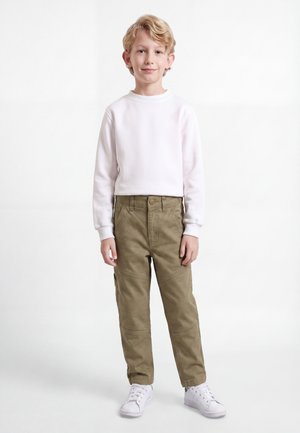 Garçon blond debout, portant une chemise blanche à manches longues, un pantalon cargo vert olive et des baskets blanches, sur un fond blanc uni.