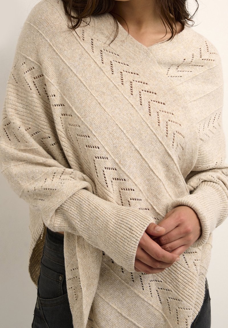 Beige wikkeltrui met een diagonaal patroon en opengehaakte accenten. Lange mouwen met ribgebreide manchetten. Zacht en warm wolmixmateriaal.
